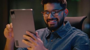 Hintli iş adamı gülümseyen Arap işadamı. Karanlık gece ofisinde dijital tablet cihazıyla konuşuyor. Video konferans konferansı konferansında sohbet görüşmesi yapıyor.