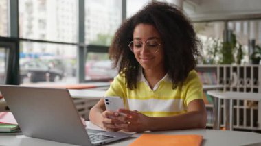 Afrikalı Amerikalı kız öğrenci zımbırtısı bağımlısı cep telefonu kullanıyor sosyal medyada konuşuyor üniversitede akıllı telefona bakıyor güzel etnik kadın sınavlarına hazırlanıyor Laptop eğitimi alıyor.