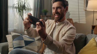 Neşeli adam, beyaz erkek, ev sahibi, oyun bağımlısı, cep telefonu bağımlısı, video oyunu oynayan, video oyunu oynayan, evinde oyun oynayan, internet oyunu oynayan, akıllı telefon esport oynayan.
