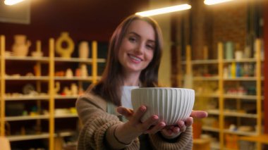 Kadın çömlekçi heykeltraş kil ustası seramik atölyesinde çalışıyor. Beyaz kadın gülümsüyor. Kameraya el yapımı bir tabak yapıyor.