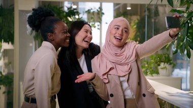 Ofisteki üç farklı çok ırklı iş kadını gülümseyen mutlu kızlar cep telefonu kamerası öğrencilerine poz veren Asyalı Müslüman Afrikalı iş kadınları iş arkadaşları neşeli kadın selfie akıllı telefon sabıkası medyası