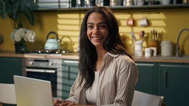 Arap Latin iş kadını etnik Hintli kız ev mutfağında dizüstü bilgisayar öğrenimi görüyor. Mutfak bayan işkadını kameraya mutlu bir şekilde gülümsüyor. İnternet üzerinden internet üzerinden alışveriş yapan yiyecek dağıtımı yapıyor.
