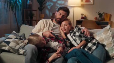 Uyuyan aile, karanlık bir oturma odasında kanepede birlikte uyur anne baba, küçük kız çocuğuyla uyur televizyon izleyip uzaktan kumandayı düşürür.