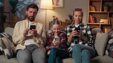Avrupalı aile cep telefonu kullanıyor akıllı telefonlar bağımlılık online internet cihazları evde akşam oturma odasında baba esneyen anne ve çocuk kız çocuk birlikte rahatlıyor