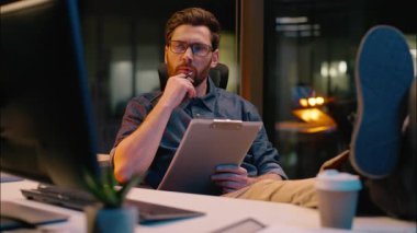 Düşünceli erkek işadamı, Kafkasyalı iş adamı, karanlık akşam ofisinde rahat bir şekilde bilgisayarlara bakıyor, notlar yazıyor evrak işleriyle uğraşıyor, beyin fırtınası fikirleri yazıyor, gece işi planlıyor.