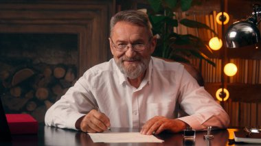 Kafkasyalı yazar elinde dolmakalem kâğıdı metin yazarı yazar kameraya bakıp gülümseyen, olgun, yaşlı, emekli iş adamı ev işleri bürosu belgelerine mektup yazıyor.