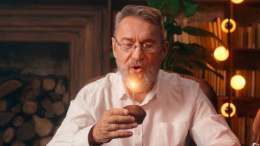 Mutlu, neşeli, yaşlı, beyaz adam mum pastası üflüyor. Doğum günü tatili. Yaşlı emekli, olgun erkek yuvasını kutluyor.