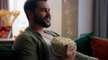 Kafkas sakallı mutlu iş adamı erkek iş adamı ev sahibi müşteri evde oturmuş patlamış mısır kasesi yiyor uzaktan kumandalı film kullanarak TV dizisi izliyor.