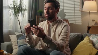 Hüsrana uğramış, üzgün, beyaz, ev sahibi, ev sahibi, hayal kırıklığına uğramış, erkek, cep telefonuyla internette geziniyor. Kanepe, akıllı telefon. Kötü haber, oturma odasındaki dolandırıcılıktan şüpheleniyor.