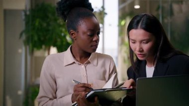 Afrikalı Asyalı iş ortakları iki farklı kız iş kadını Çinli Amerikalı iş arkadaşları gülümseyen kadın takım çalışanları ofis yöneticileri dizüstü bilgisayar projelerini tartışıyor notlar yazıyorlar