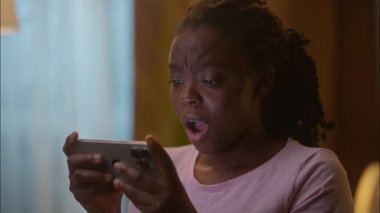 Korkmuş Afrikalı Amerikalı etnik iş kadını kadın kullanıcı kız elinde cep telefonu ile oyun oynuyor bahis kaybediyor ev yatak odasında başarısız cep telefonu hüsranı