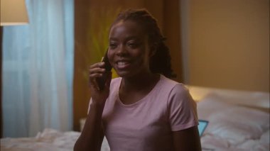 Neşeli kahkahalar atan Afro-Amerikan etnik kadın öğrenci kız serbest çalışan cep telefonuyla konuşan, akıllı telefon konuşabilen sohbet eden komik hikaye anlatan ev yatak odasında geç saatlere kadar çalışan dizüstü bilgisayar.