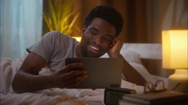 Kahkahalar atan, Afro-Amerikalı, erkek, ev sahibi, kullanıcı, elinde tablet olan bir müşteri, gece yarısı yatak odasında sosyal medya komedisi seyrediyor.