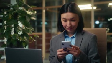 Asyalı Çinli Japon kadın iş kadını elinde cep telefonuyla akıllı sohbetler internette mesajlaşmalar dizüstü bilgisayarlı ofis çalışanı bayan işveren kız işyeri müdürü