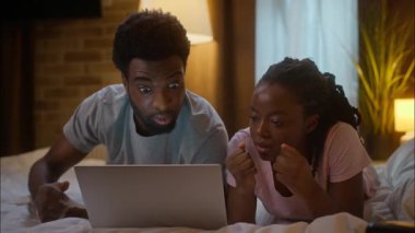 Üzüntülü Afrikalı Amerikalı aile eşi çift yatağında yatıp dizüstü bilgisayar seyrediyor hayal kırıklığına uğramış taraftarlar için tezahürat yapıyor ev yatak odasında kaybedilen maçı spor takımı kaybediyor