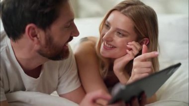 Kafkasyalı mutlu erkek, sabah yatağında yatan erkek, tablet yazan kadın, sosyal medya eşiyle sohbet ediyor. İnternet üzerinden alışveriş yapıyorlar. Yatak odasında gülümseyen eşler.