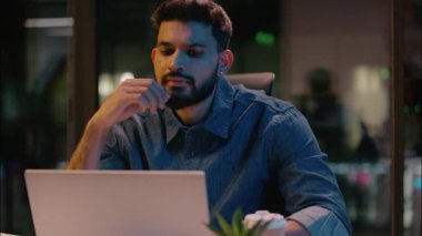Gece vardiyasına kalan ofis işleri Hintli Arap etnik adam erkek, erkek girişimci iş adamı iş yerinde iş fikri yaratmayı düşünüyor bilgisayarın düşünceli iç görü planına ilham veriyor.