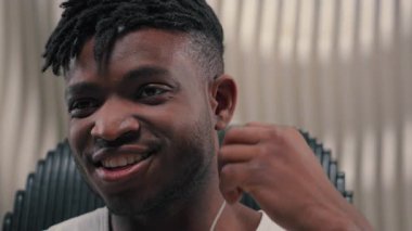 Afrikalı Amerikalı siber adam gülümsüyor, telli kulaklık takıyor, müzik dinliyor, iyi ses kalitesinde kulaklıklar takıyor, beyaz fütürizm stüdyosundaki kulaklıklardan keyif alıyor, içeride mutlu fütüristik teknoloji.