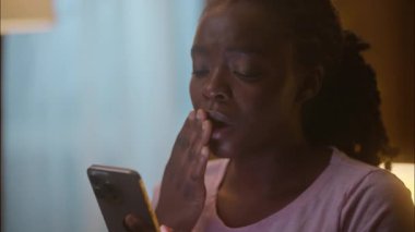 Uykulu, yorgun, Afrikalı, Amerikalı kadın, bayan, müşteri müşterisi, cep telefonu kullanıyor. Sosyal medyada kayıyor. Uyku aleti bağımlılığından önce uykusuzluk çekiyor.