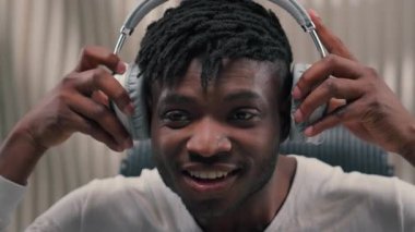Mutlu genç Afrikalı Amerikalı erkek kulaklıklarını takmış içeride müzik dinliyor ritmin tadını çıkarıyor fütüristik oda fütürizminde neşeli duygular hissediyor.