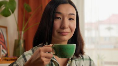 Asyalı Koreli Çinli kadın Japon kadın portresi aromalı çay içiyor bitkisel kahve içiyor cappuccino latte espresso içiyor elinde fincanı tutuyor ve sabah kahvaltısında kameraya bakıp gülümsüyor.