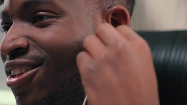 Afrika kökenli, mutlu, gülümseyen bir adam. Telli kulaklık takıyor, dinleniyor, müzik dinliyor, beyaz fütürizm odasında kaygısız bir şekilde şarkı dinliyor.