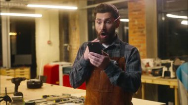 Kafkasyalı mutlu beyaz adam ahşap ustası atölye marangozunda cep telefonu kaydırma başarısını kutluyor. İşçi erkek cep telefonu tamircisi neşeli internet uygulamaları kullanıyor.