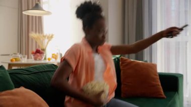 Mutlu, neşeli, gülümseyen, dağınık, Afro-Amerikalı, iş kadını, bayan, iş kadını, ev sahibi, hafta sonu rahatlığı, elinde patlamış mısır kasesi, evinde televizyon izleyen bir kontrolör.