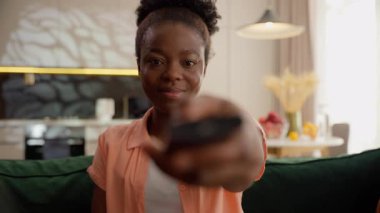 Mutlu gülümseyen Afrikalı Amerikalı etnik iş kadını bayan iş kadını kız izleyici kanal değiştirici uzaktan kumanda TV televizyon dizisi filmi izlerken ev kanepede patlamış mısır yiyor.