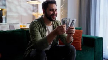 Mutlu gülümseyen beyaz iş adamı erkek müşteri ev sahibi zengin iş adamı serbest girişimci kanepeye oturmuş dolar tutan nakit para kazanan zafer evde kumar oynayarak lotoyu kazanıyor.