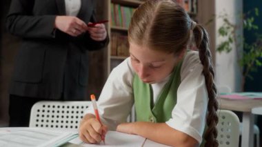 Beyaz kadın öğretmen, gülümseyen profesör öğrencinin hata düzeltmesine yardım ediyor. Küçük kız çocuğuna kalem ödevi yazmasını öğretiyor.