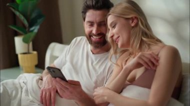 Gülümseyen beyaz aile çifti yatak odasında cep telefonu kullanırken mutlu kadın cep telefonunu kaydıran adam sosyal medya görüntüsü veren internet kocası karı koca sohbeti sabah internetten alışveriş yapıyor.