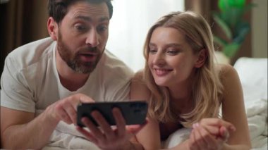 Neşeli Kafkasyalı çift cep telefonunu ev arkadaşının cep telefonunda kullanıyor internetten birlikte ağ satın alıyorlar olumlu adam internetteki internet sitesine kayıyor internetteki uygulamalı kadın gülüyor yatak aşkı sabahı