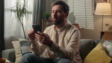 Üzgün, hayal kırıklığına uğramış, Kafkasyalı işadamı koltukta cep telefonu kullanmaktan sıkılmış, hayal kırıklığına uğramış, erkek ev sahibi evde cep telefonu okuyup mesajları karıştıran akıllı telefon.