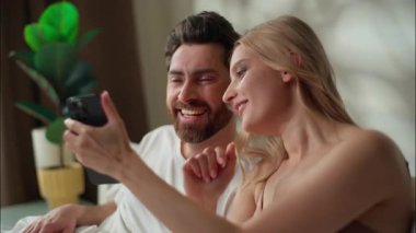Mutlu Kafkasyalı çift cep telefonunu yatak odasında birlikte kullanıyor erkek arkadaş gülüyor akıllı telefon kadını internet üzerinden alışveriş yapan internet uygulamaları alıyor.