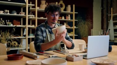 Beyaz tencereli çömlekçi elinde çanak çanak çömlek ürünü porselen el işi atölyede gülümseyen erkek heykeltraş tasarımcı seramik sanat eserleri gösteren küçük işletme sahibi.