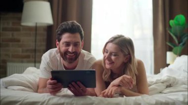 Kahkaha atan beyaz bir aile yatak odasında tablet tutan evli bir çift neşeli sevgili yazıcı kadın internetten alışveriş yapan internetçi adamla sabah siber-uzay alışverişi yapıyor.