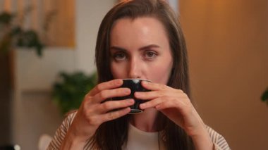 Beyaz kadın gülümsüyor sıcak aromalı çay içiyor. Kahve fincanı tutan iş kadını kafeteryada oturmuş kameralı kafeterya ziyaretçisine bakıyor. Kahvaltının keyfini çıkarıyor.