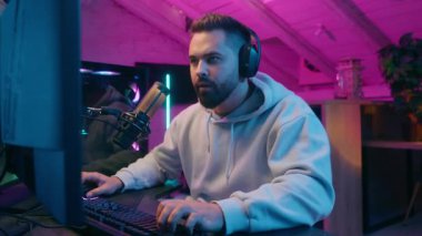 Bağımlı Kafkasyalı erkek oyuncu erkek oyuncu flamalı online savaş oynuyor. Kulaklık takarak gülüyor, bilgisayarlarda oyun oynuyor. Evdeki karanlık odada dijital oyun oynuyor.