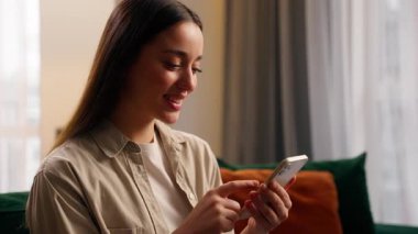 Beyaz kadın evde cep telefonu kullanıyor mutlu kız sohbet ediyor oturma odasında akıllı telefon ile sohbet ediyor internetteki uygulama sosyal medyada komik videolar seyrediyor internet alışverişini tatmin ediyor.