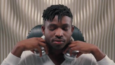 Afro-Amerikalı erkek, kulaklık takıyor, müzik dinliyor, sandalyeye yaslanıyor, keyfine bakıyor, gelecekteki oda fütürizm stüdyosundaki kulaklıklardan müzik dinliyor.