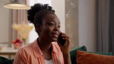 Ciddi Afrikalı Amerikalı iş kadını kadın etnik bayan iş kadını ev sahibi model müşteri cep telefonuyla konuşuyor cep telefonuyla konuşuyor kapalı bir evde koltukta oturuyor.
