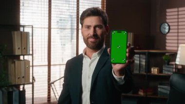 Gülümseyen orta yaşlı iş adamı, yeşil ekranlı cep telefonunu işaret eder gibi gösteriyor. Akıllı telefon kromu, anahtar gösterim başarısı. Ofiste kameraya bakan olgun bir iş adamı elde et.