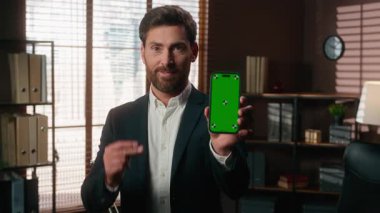 Gülümseyen orta yaşlı iş adamı akıllı telefon kromasını işaret eden yeşil ekranlı bir cep telefonu gösteriyor. Kumarhane uygulaması yetişkin bir iş adamı ofisinde kameraya bakıyor.