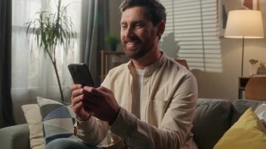 Mutlu adam cep telefonu kullanıyor gülümsüyor Kafkasyalı erkek iş adamı akıllı telefonuna bakıyor iyi haber mesajı neşeli ev sahibi oturma odasındaki kanepede dinleniyor online alışveriş uygulaması teslimatı