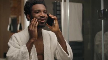 Banyodaki Afrikalı Amerikalı adam sabah duştan sonra cep telefonuyla konuşuyor sakalını kesiyor gülümseyen sakallı adama dokunuyor arkadaşça sohbet ediyor otel banyosunda cep telefonuyla iletişim kuruyor.