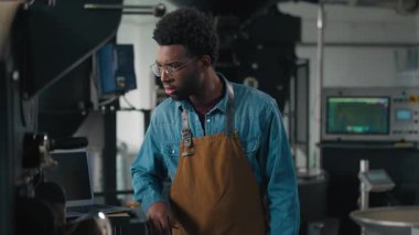Afrika kökenli Amerikalı bir adam endüstriyel kızartma makinesi kullanarak kahve çekirdeği kızartıyordu. Önlüklü, kafası karışmış erkek kızartma makinesini kontrol ediyordu. Ellerini çapraz yapıp modern fabrika çalışanı baristaya gülümsüyordu.