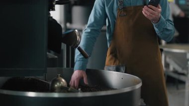 Kafkasyalı bir adam kavrulmuş kahve çekirdeklerini teftiş ediyor kızartma makinesinde kontrol ediyor mesaj niteliğinde verileri çevrimiçi hizmet veren cep telefonu erkek kafein dağıtımında yazıyor.