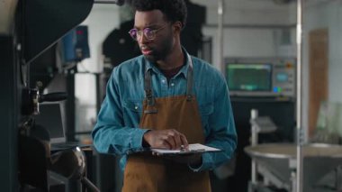 Afrika kökenli Amerikalı bir adam kavrulmuş kahve fabrikası mühendisi. Endüstriyel kızartma makinesinin bazı kısımlarını kontrol ediyor. Yazı yazıları yazıyor.