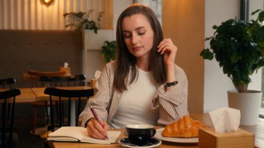 Kafedeki beyaz kadın not defterine notlar yazıp kahve içerken kafeteryadaki mutlu bayan kız çay içerken kruvasanla günlük bilgileri yazıyor.